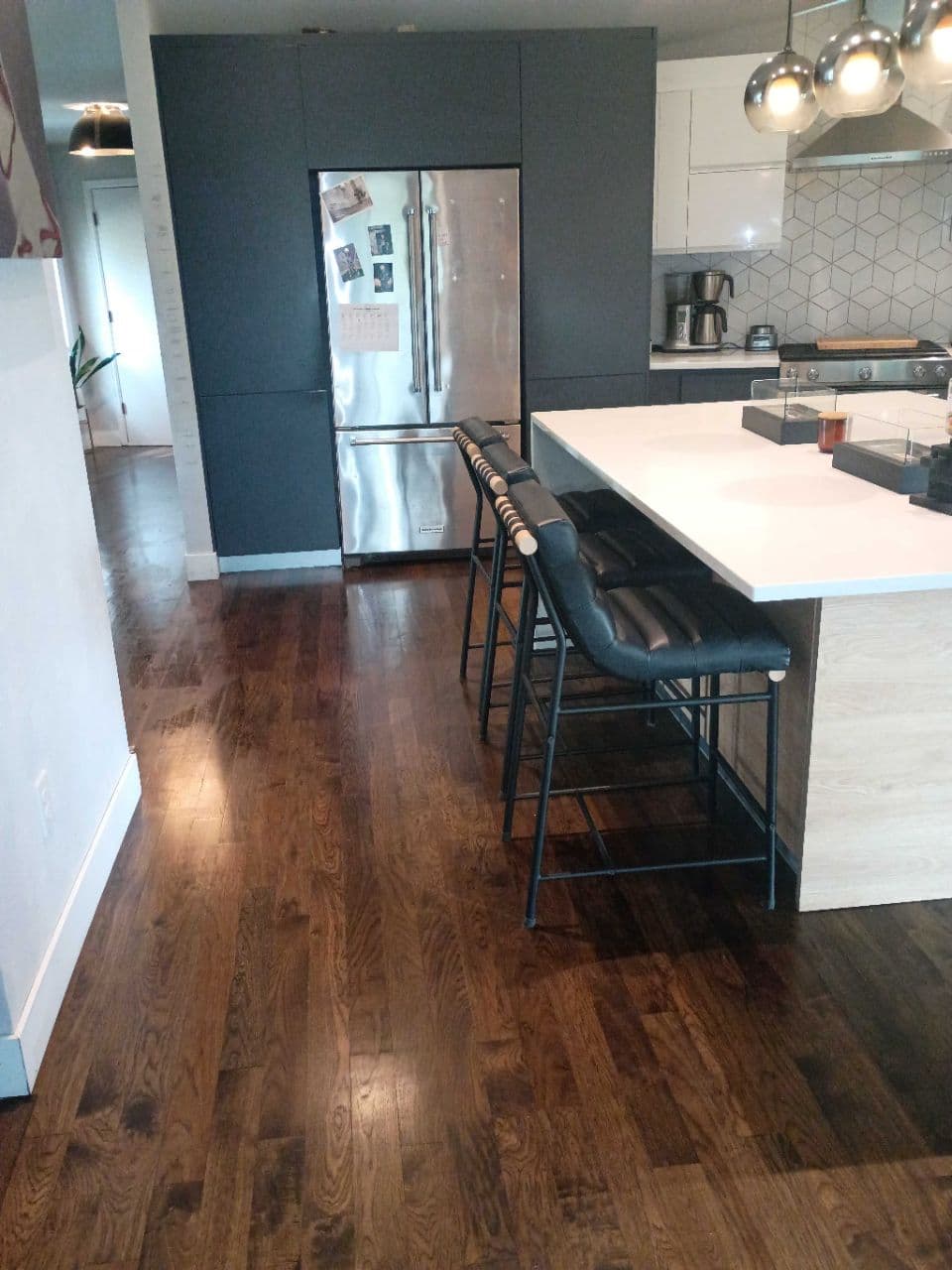 Modern kitchen with dark hardwood floors, white island, black bar stools, and stainless steel refrigerator.