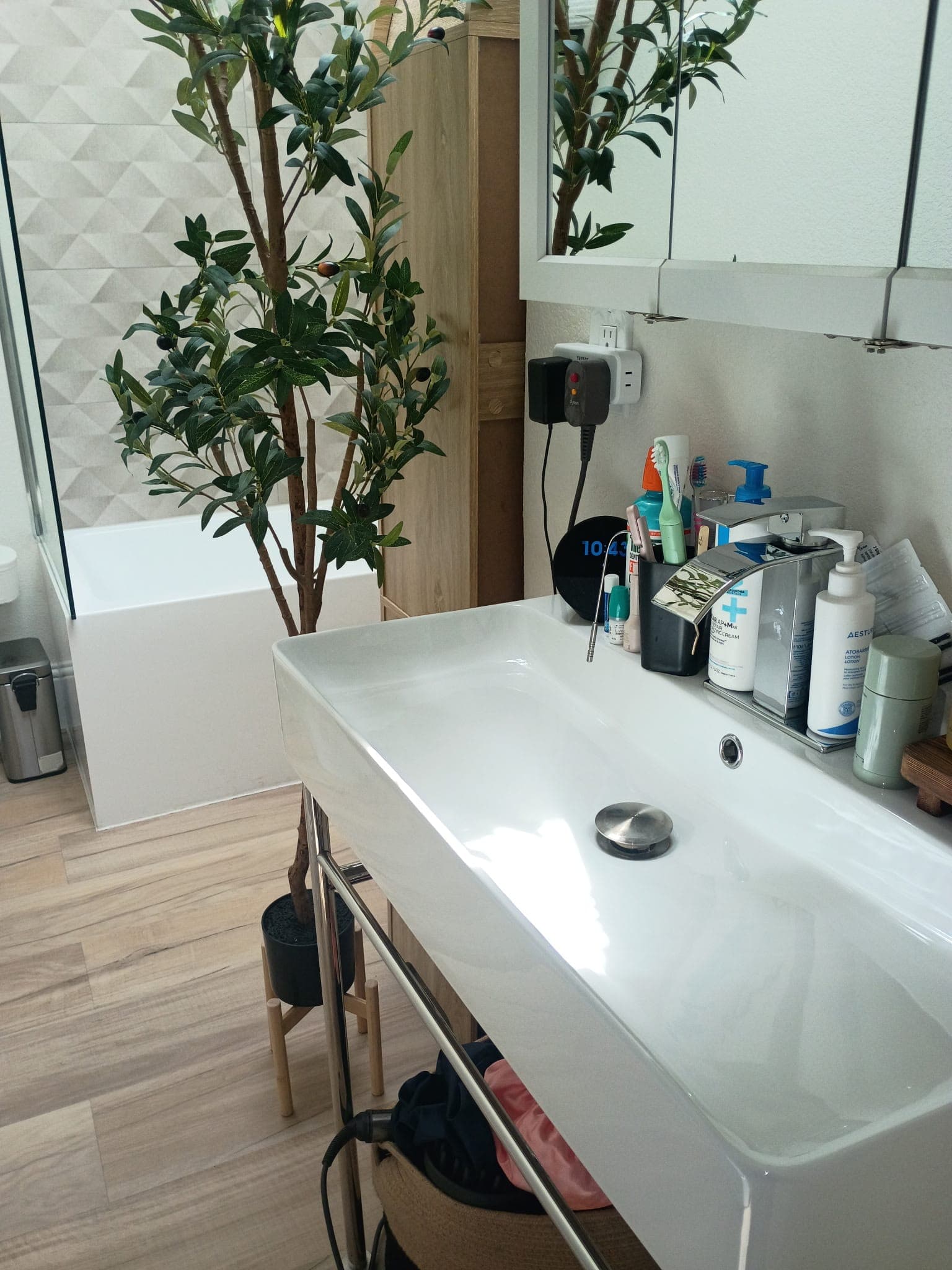 White rectangular bathroom sink with toiletries, chrome faucet, and a tall potted olive tree.